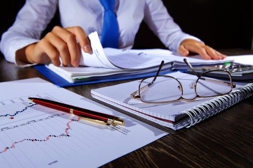Scrivania con fogli e occhiali e un uomo seduto che sfoglia delle carte